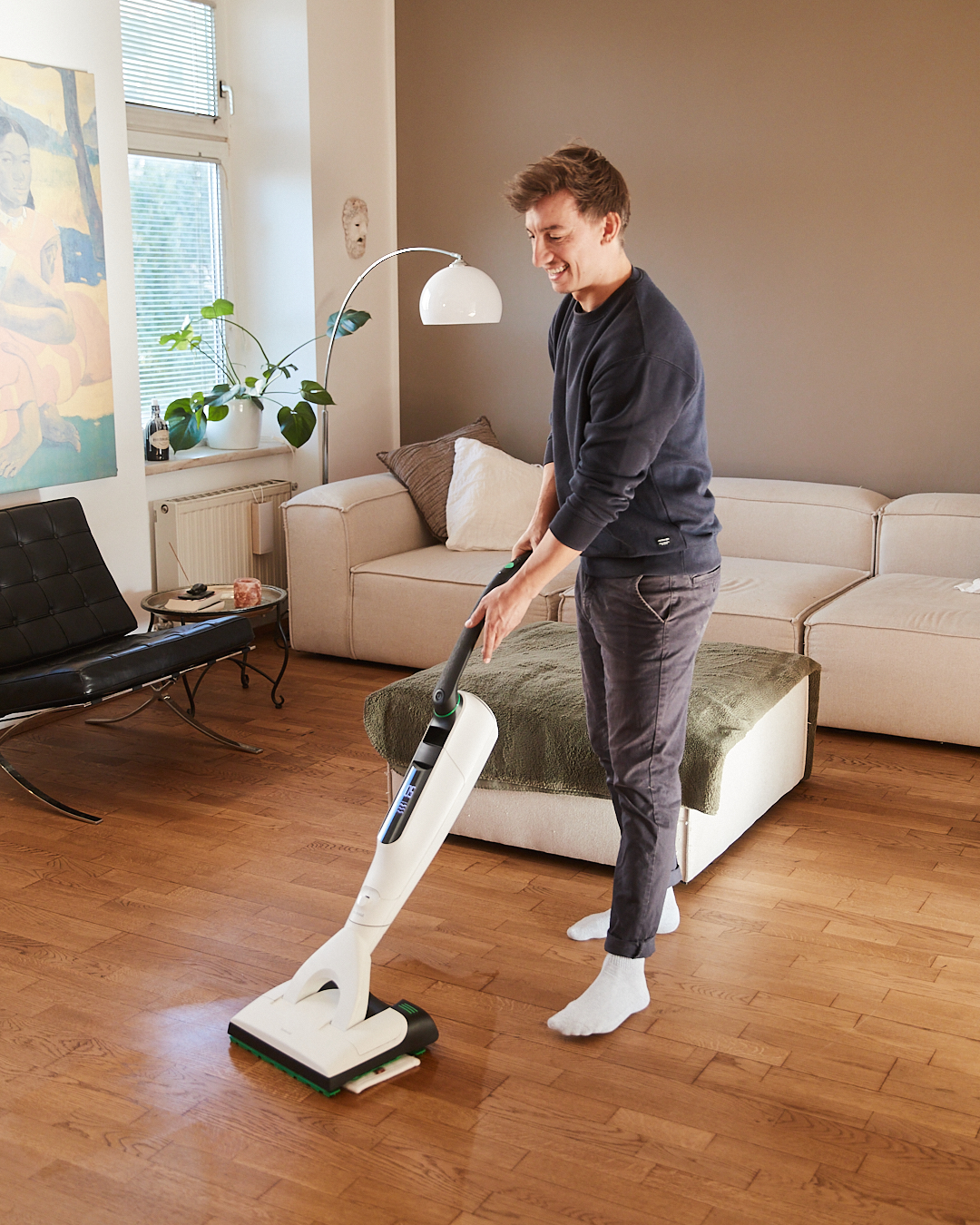 Mann wischt lächelnd den Holzboden im Wohnzimmer mit dem Kobold SP7 – Szene aus der aime Kampagne „Mental (un)load“ für Vorwerk.